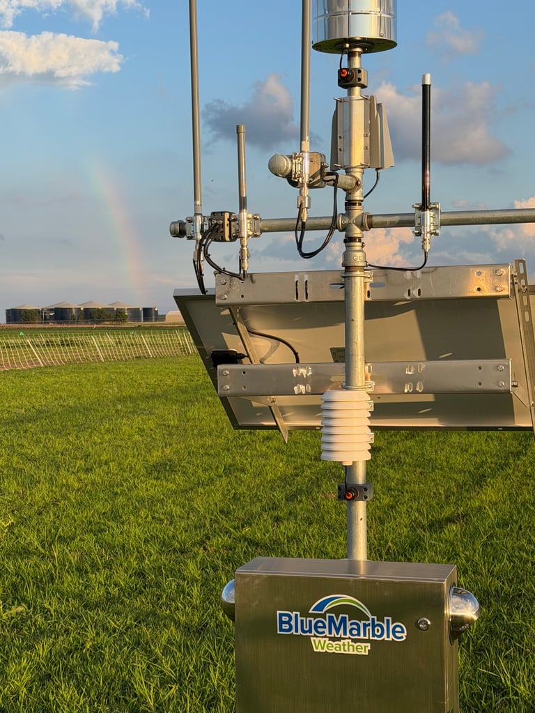 BlueMarble Weather station with multiple antennas and sensors mounted on a pole in a green farm field under blue sky